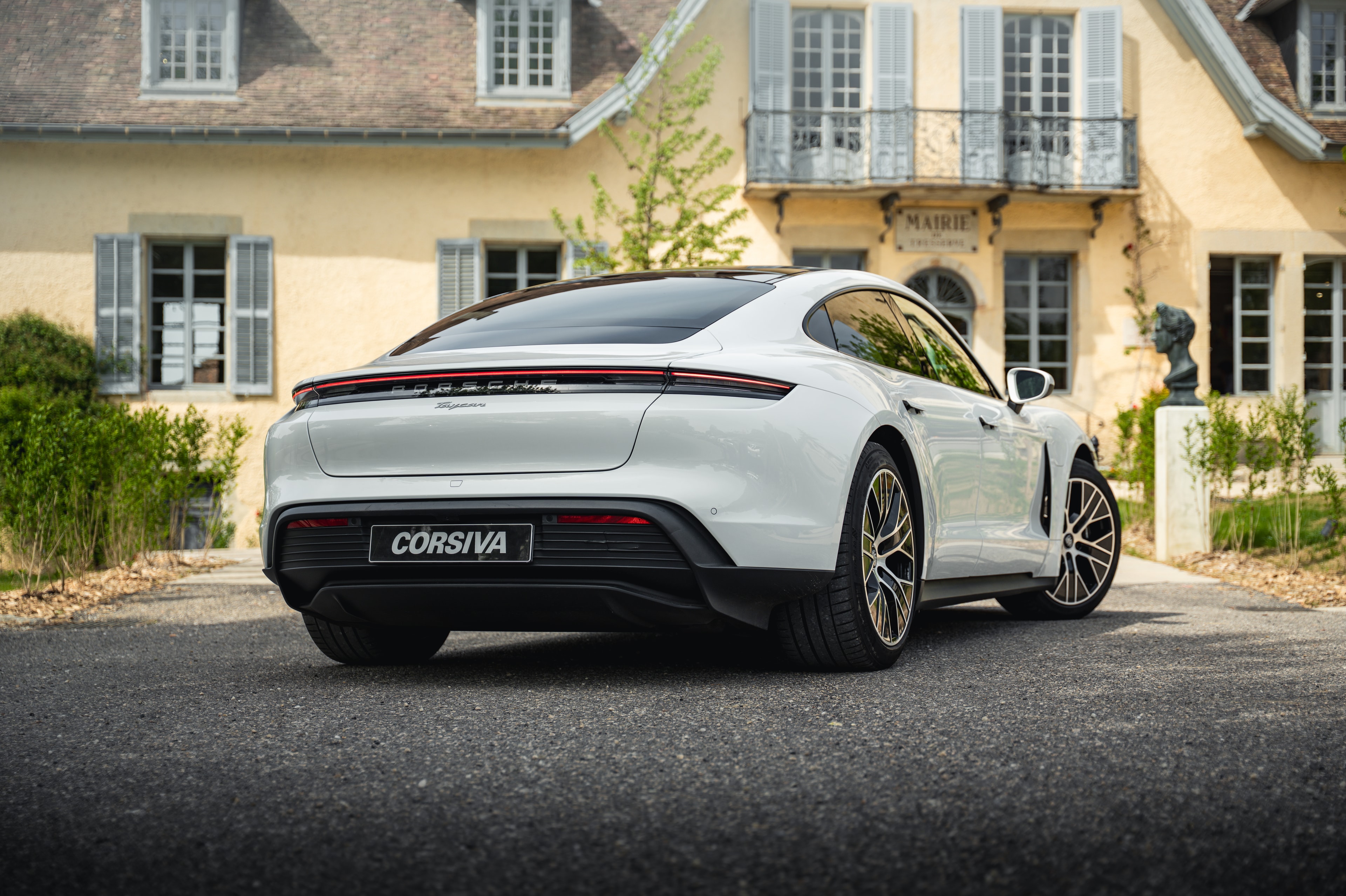 Porsche Taycan blanche avec plaque Corsiva, vue de trois quarts arrière devant la façade d’une demeure de caractère